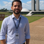 Pessoa em primeiro plano usando camisa social azul-clara e crachá pendurado no pescoço, posicionada em uma área aberta da Esplanada dos Ministérios, em Brasília. Ao fundo aparecem os prédios do Congresso Nacional, gramados verdes, céu azul com nuvens e uma placa com o texto “Esplanada dos Ministérios”. A imagem foi registrada durante o dia, com iluminação natural e boa visibilidade dos elementos arquitetônicos ao redor.