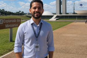 Pessoa em primeiro plano usando camisa social azul-clara e crachá pendurado no pescoço, posicionada em uma área aberta da Esplanada dos Ministérios, em Brasília. Ao fundo aparecem os prédios do Congresso Nacional, gramados verdes, céu azul com nuvens e uma placa com o texto “Esplanada dos Ministérios”. A imagem foi registrada durante o dia, com iluminação natural e boa visibilidade dos elementos arquitetônicos ao redor.